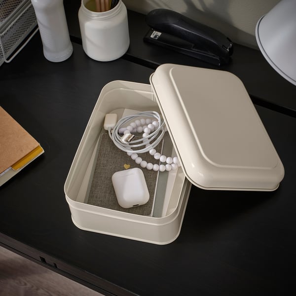 A beige metal box on a dark desk containing a white mouse, a white charger cord, and a white stylus. Used for organising small items.