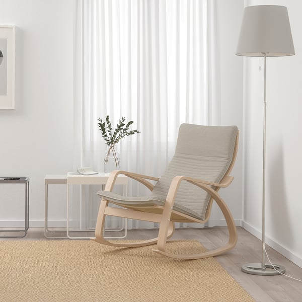 Minimalist living room with POÄNG chair, footstool, side table, plants, and lamp. Light wood chair near window with sheer curtains and large rug.