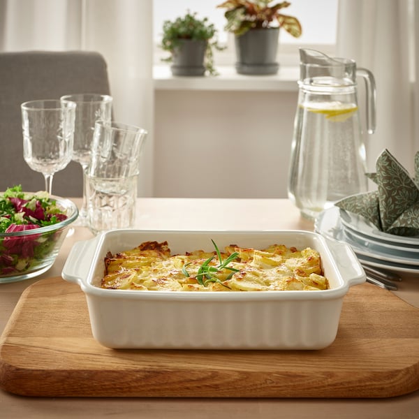 White stoneware gratin on wooden board, with glasses, pitcher, and salad bowl on bright table.