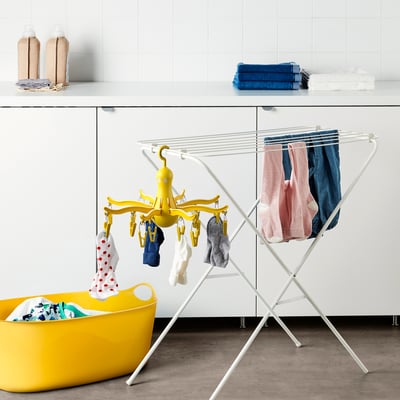 Bright laundry scene: yellow PRESSA dryer hangs small clothes; next to white cabinet, yellow laundry basket, blue towels, two bottles.