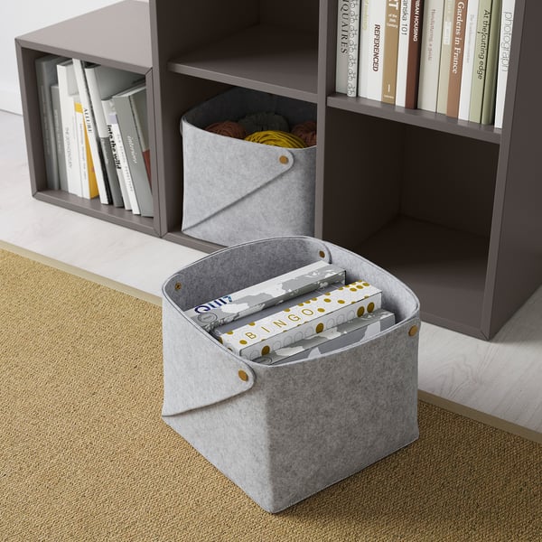 Two grey fabric storage cubes, one holding boxes, the other yarn, beside a grey bookshelf with books.