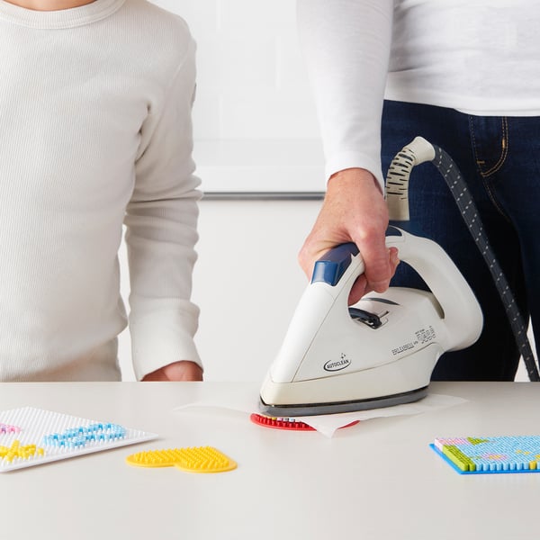 A person irons a fused bead project. Yellow, blue, pink beads are on a white table. Kmart iron adheres them.