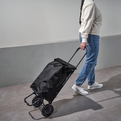 Person pulls black RADARBULLE foldable shopping cart with adjustable handle and large bag on concrete surface.