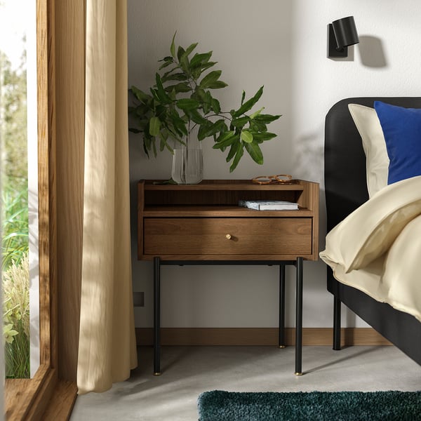Bedroom scene featuring mid-century RÅDMANSÖ bedside table in warm walnut with black metal legs, cream curtains, and a blue pillow on a bed.