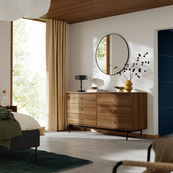 A modern bedroom a RÅDMANSÖ chest with six walnut drawers and gold knobs, set against a white wall with a round mirror and yellow vase with flowers.