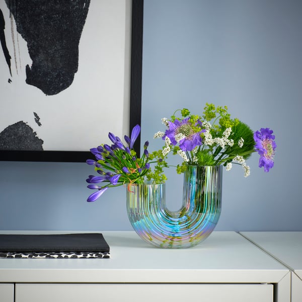 A rainbow-like RÄFFELBJÖRK vase with purple flowers, on a white table beside a book and picture.