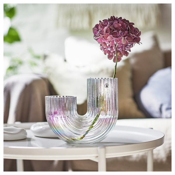 A clear, rainbow-inspired clear glass vase named RÄFFELBJÖRK by lisa hilland, filled with a bright purple flower, sitting on a white table.
