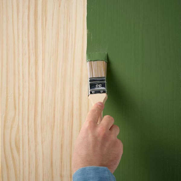 A hand holds a paintbrush applying green paint to a wooden surface, demonstrating the woods paintability for customization.