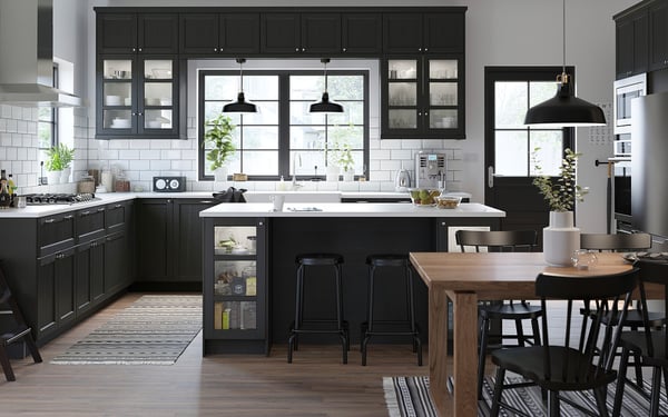 Modern kitchen with black cabinets, white counters, glass cabinets, island, black stools, large windows, pendant lights, black & white rug, plants.