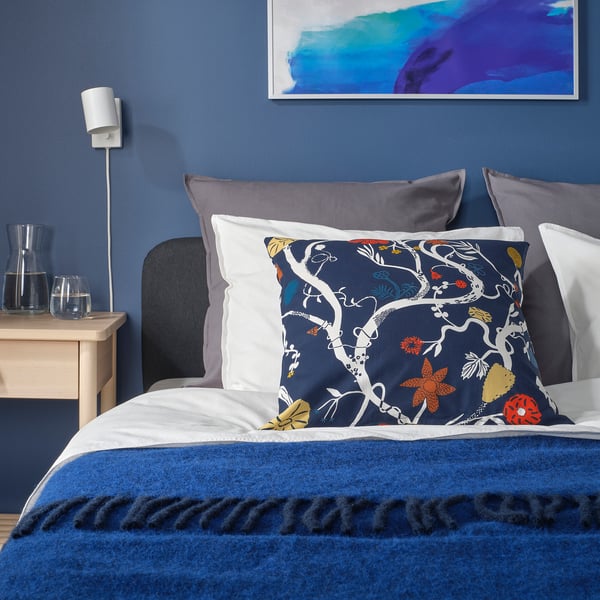 Navy blue REVFINGERÖRT floral cushion on stylish bed with mix pillows, throw, and wooden bedside table.