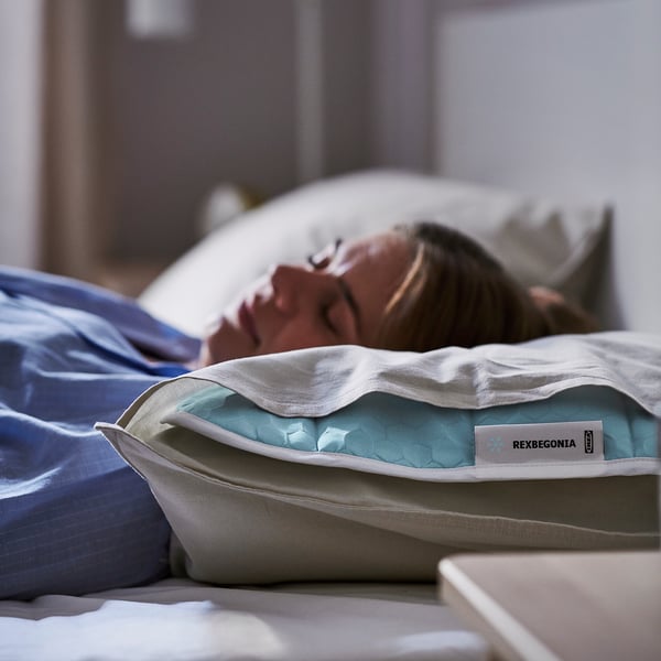 A person napping on a bed with a REXBEGONIA blue cooling pad supporting them. The pad has a white zip and tag.