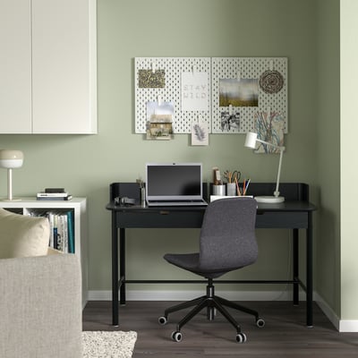 Black desk and chair in green corner with laptop and pegboard.