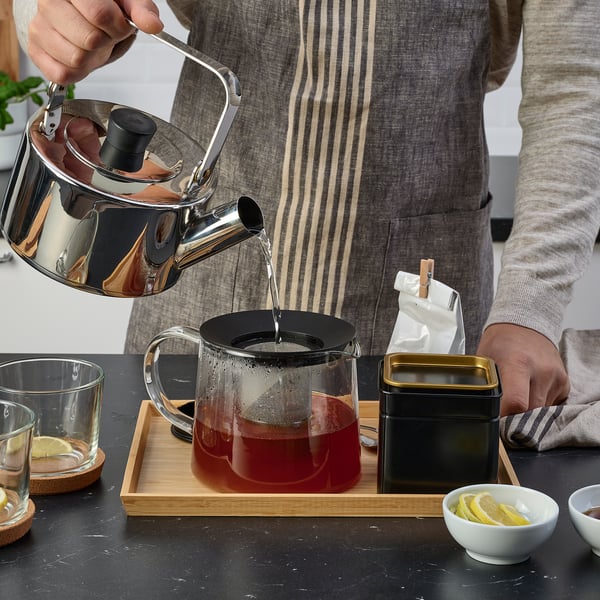 Pouring tea into glass teapot with infuser on wooden tray, surrounded by cups, lemons, and tin.
