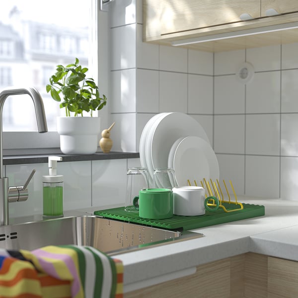 Kitchen sink with green dish drainer holding plates, cups, and cutlery. Beside is soap dispenser and plant.