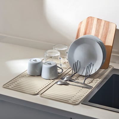 Kitchen counter with RINNIG mat holding organised dishes and utensils, near wooden cutting board.