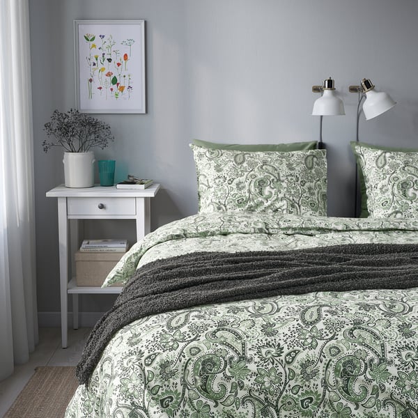 Cosy bed with green paisley bedding, geometric lamps, small white side table holding books & vase, light grey walls, art, & sheer curtains.