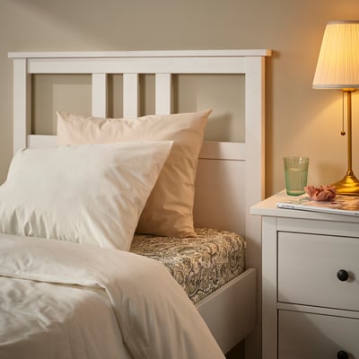 A cosy white bed with beige and white pillows, a small green glass, a lamp, and a book on a white bedside table beside it.