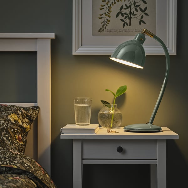 Cosy bedroom with green RÖDFLIK desk lamp on small table beside bed, featuring adjustable shade, glass of water, and small plant vase.