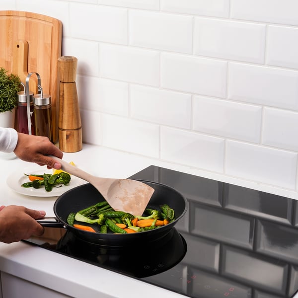 Person cooks vegetables in a skillet on a white stove. Wooden spatula and plate of vegetables nearby.