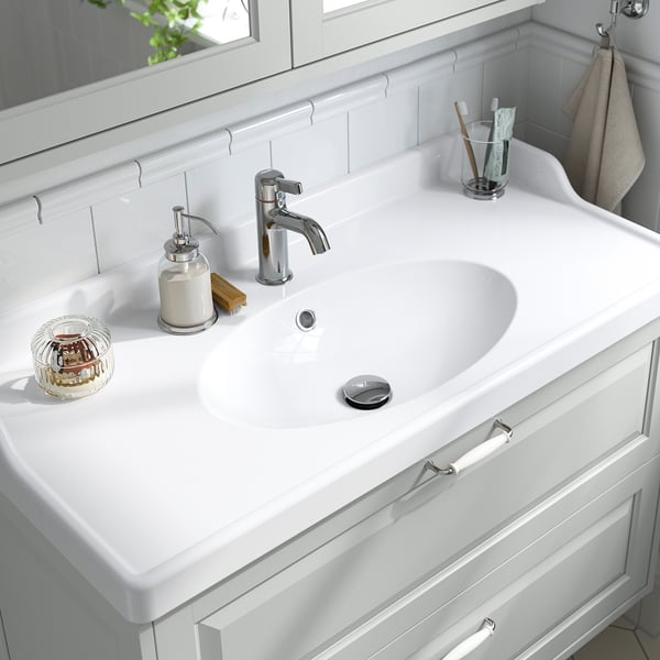 A sleek white bathroom sink with a raised backsplash, featuring a chrome tap and accessories, designed to fit seamlessly into a washstand.