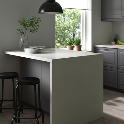 Modern kitchen island with white rounded edges, green base, two black stools, light fixture, grey cabinets, and wooden floor.
