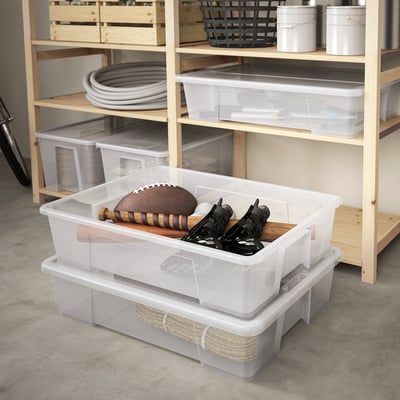 Clear plastic SAMLA boxes storing sports gear, football, skates. Top box smaller, neat setup with striped mat. Wooden shelves hold more containers.