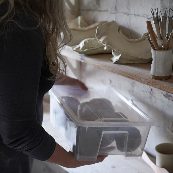 Woman holding clear plastic box with black contents. Box has built-in click lock. Wooden shelf with tools in the background.