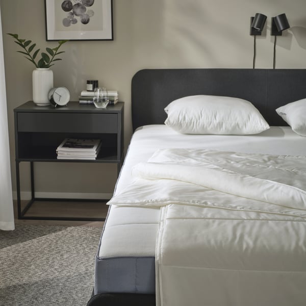 Bedroom scene with SANDGRÄSMAL duvet on bed, gray nightstand holding books, clock, and plant; black headboard, wall lamps.