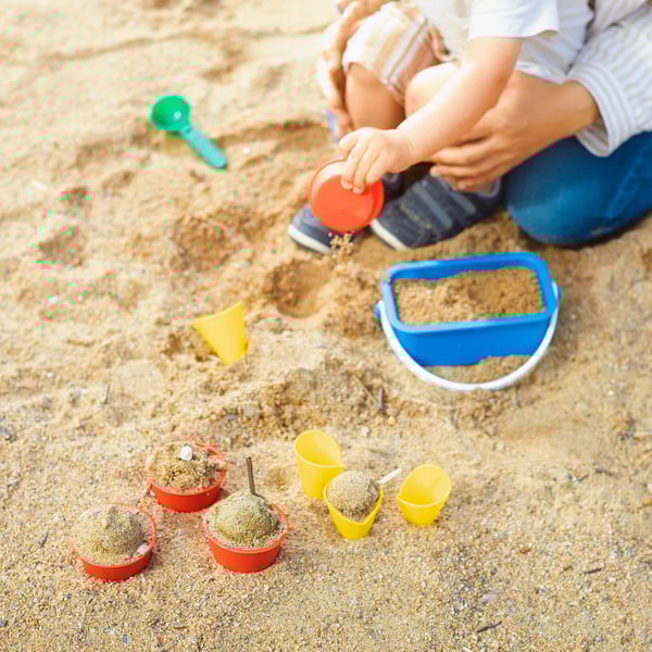 Child playing with SANDIG sand toys, including cups and a scoop, developing motor skills on the sand.