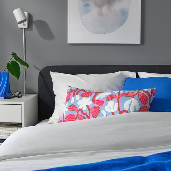 Modern bed with blue & red floral pillow, white sheets. Plant & book on side table with lamp.