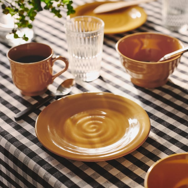 SANDSKÄDDA dinnerware in ochre yellow, including plates, cups, and a glass. Stoneware with rustic feel, stacked on chequered tablecloth.