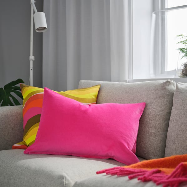 Bright pink cushion on grey couch, larger than nearby multicoloured stripes pillow, soft cotton velvet.