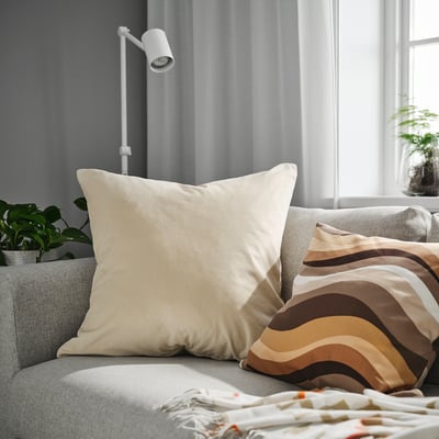 A large beige SANELA cushion cover on a grey couch, with striped pillows and plants nearby.