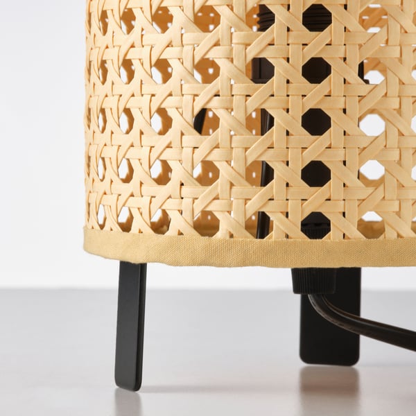Wicker-like lamp shade with intricate patterns, sitting on sleek black legs, emitting warm light.