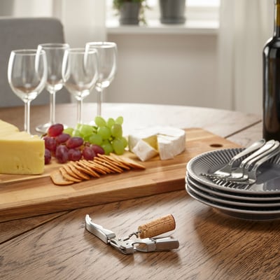 Wooden table set with cheese, grapes, crackers, wine glasses, plates, forks, a corkscrew, and a bottle, ready for a meal.