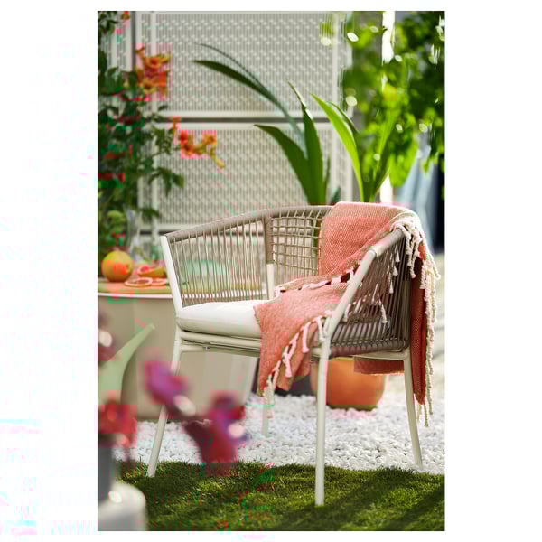 Cosy white wicker chair with orange blanket on patio, surrounded by lush greenery and flowers.