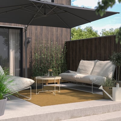 Modern patio with black parasol over white sofa on yellow rug, surrounded by potted plants and a wooden fence with greenery.