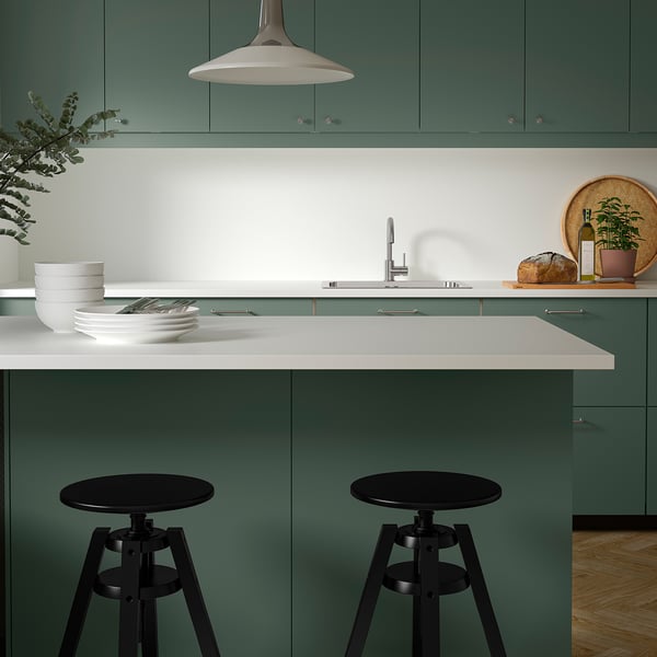 Modern teal green kitchen with sleek countertops, black bar stools, white bowls, plant, and hanging lamp.