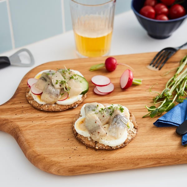 Wooden board holds two open sandwiches topped with herring, sliced vegetables, and greens. Glass of yellow drink, knife, and radishes nearby.
