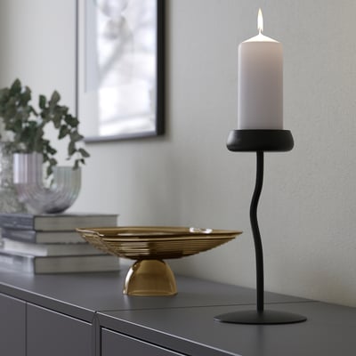 A modern black candlestick holds a white lit candle on a dark surface, offering mood lighting.
