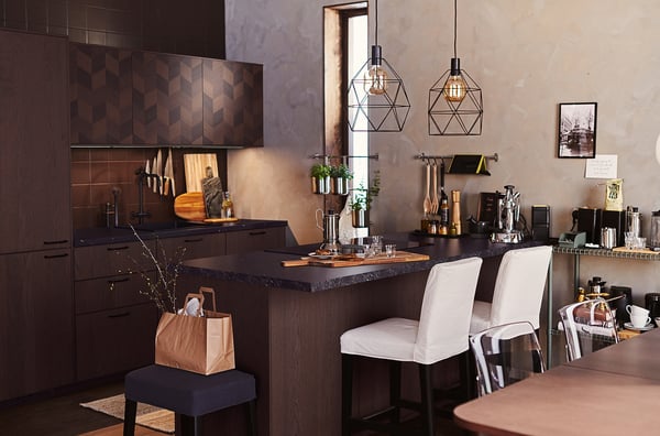 Modern kitchen with dark wood cabinets, black countertop, white chairs, geometric pendant lights, and various utensils and plants.