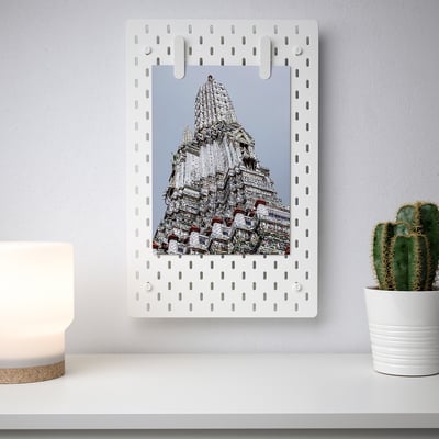 White SKÅDIS pegboard displays large photo, surrounded by evenly spaced holes. Mounted on a light wall, with a cactus plant and a lamp nearby.