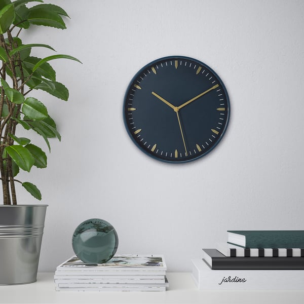 Wall clock with dark blue face and gold hands, silent quartz movement, above table with books and plant.