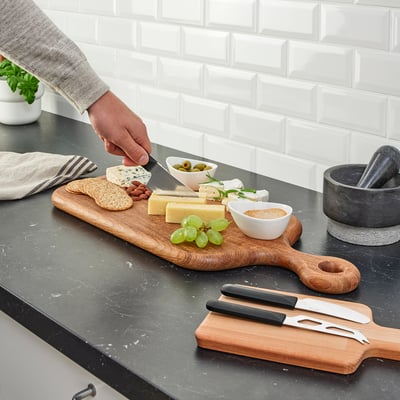 A person is cutting cheese on a wooden board with SKÄRLÅNGA knives. The board holds grapes, olives, and various cheeses.