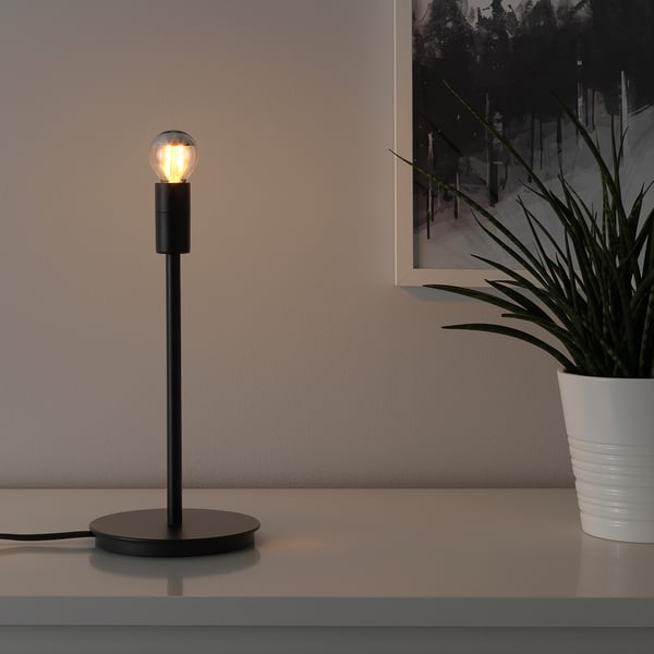 A sleek, black table lamp with a globe light bulb emits warm light on a surface next to a potted plant.
