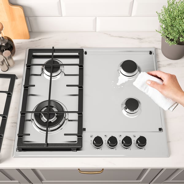 Person cleans modern gas stovetop with white cloth. Stainless steel finish, four burners, electronic ignition.