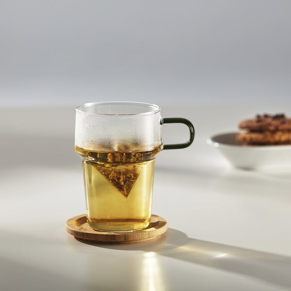 Clear glass mug with green handle holds a tea bag, placed on wooden coaster, sitting on table with sunlight reflecting off it.