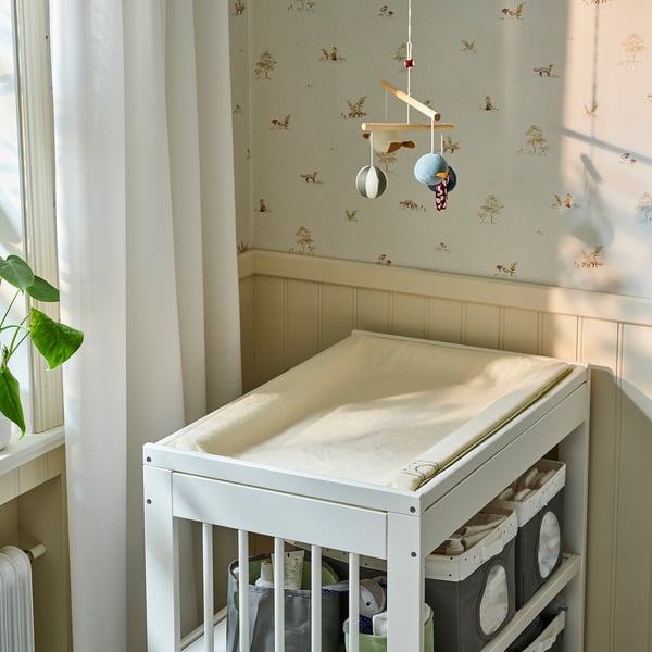 Small white ikea SKÖTSAM changing mat with cat print, compact under a mobile, next to a window.