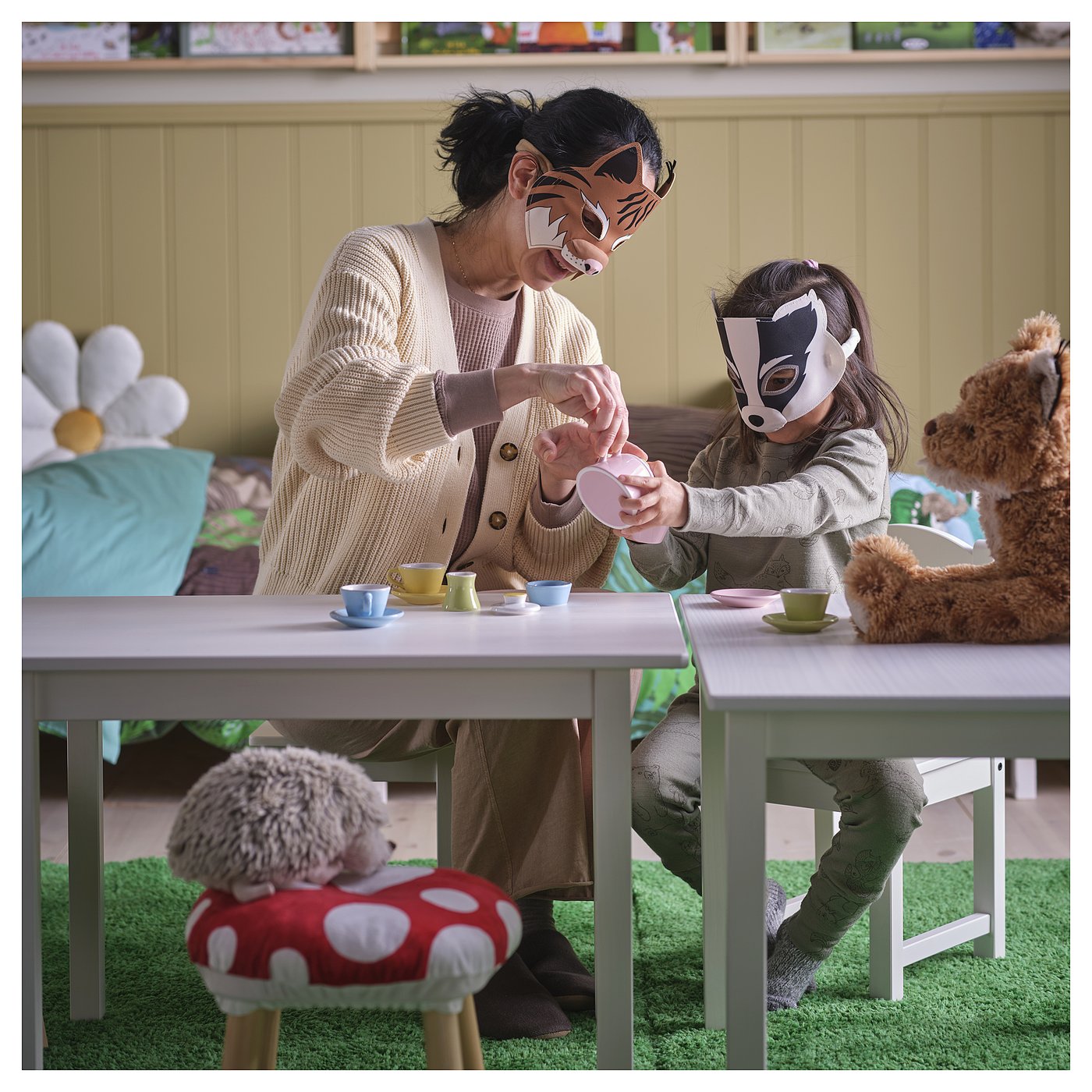 An an adult and child playing with SKOGSDUVA masks, sitting at a small table with tea cups, surrounded by stuffed animal toys.