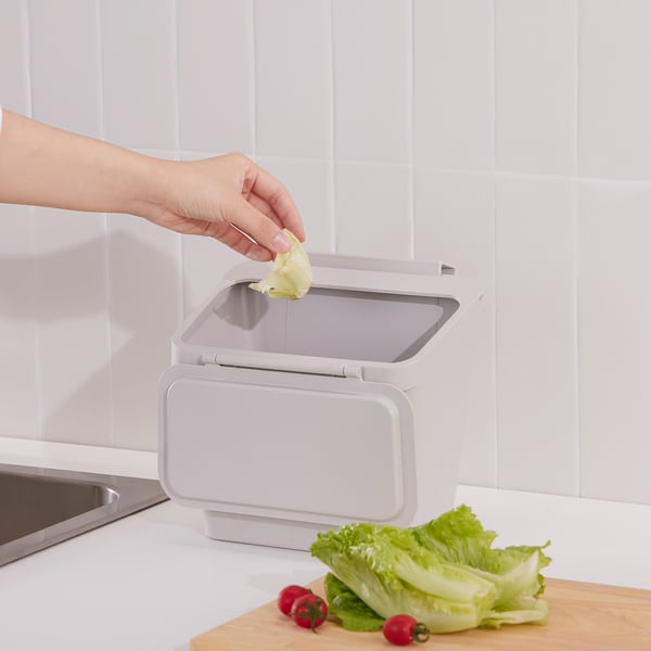Hand placing lettuce peel into SKOLÄST kitchen waste bin with lid open, set on counter near sink.